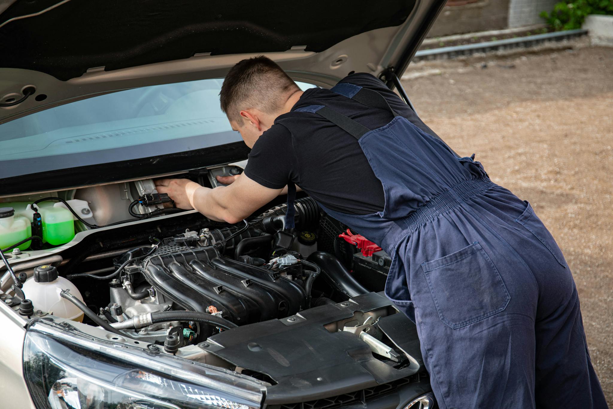 Mechanic in overalls fixing a car engine outdoors, showcasing automotive repair skills.