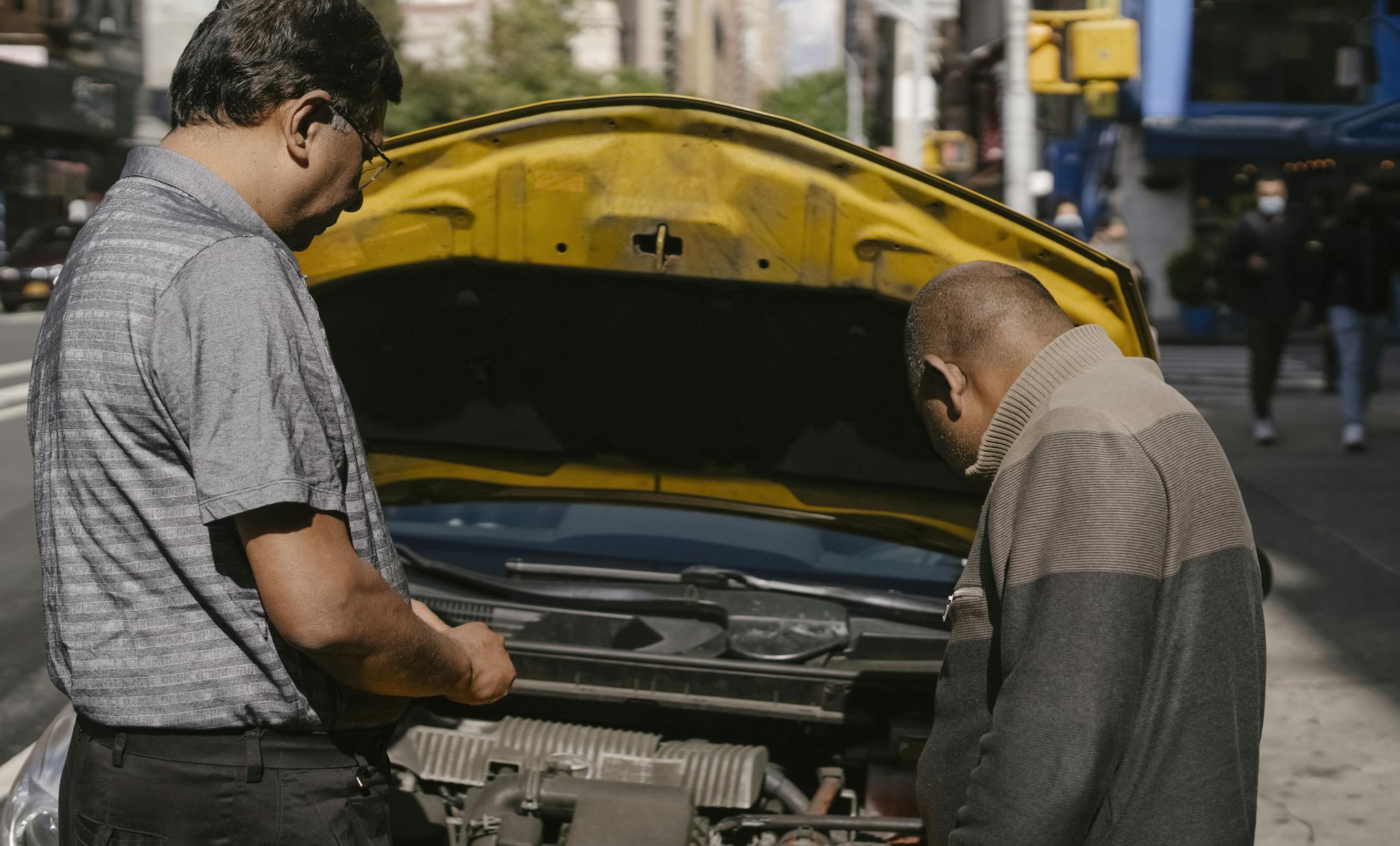 Two men inspecting a car engine on a city street, focusing on vehicle maintenance and repair.